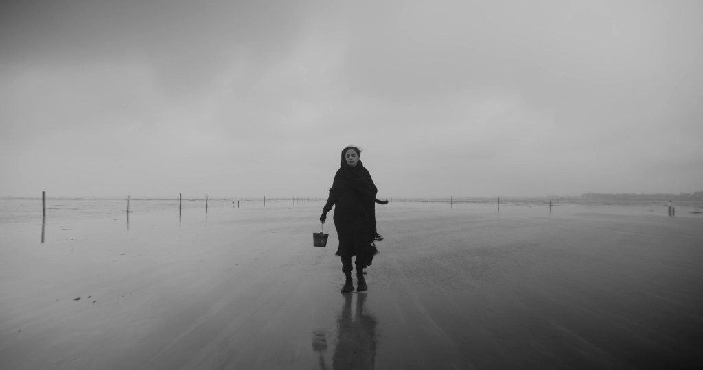 A woman in a dark shawl walks through shallow water carrying a bucket, under a grey sky. From A Drop at a Time (2025), directed by Mazyar Mahan.