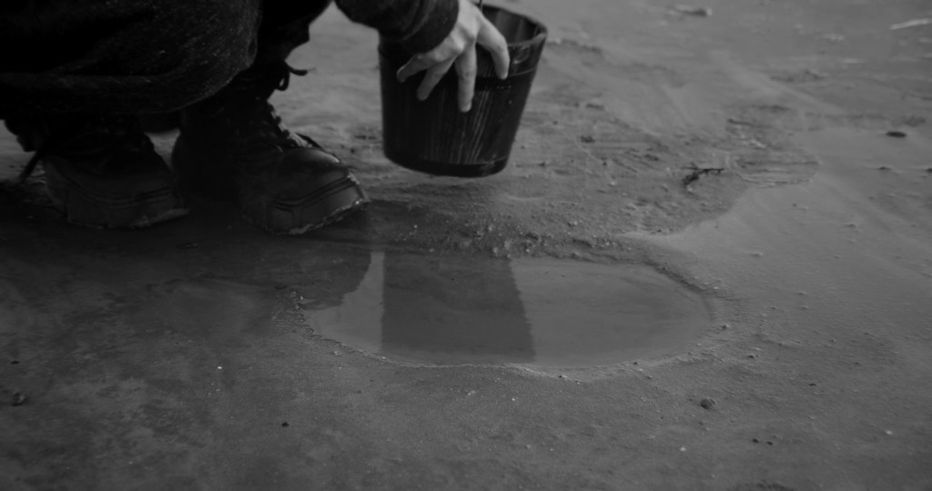 Close-up of a hand dipping a small bucket into a shallow puddle on wet sand. From A Drop at a Time (2025), directed by Mazyar Mahan.