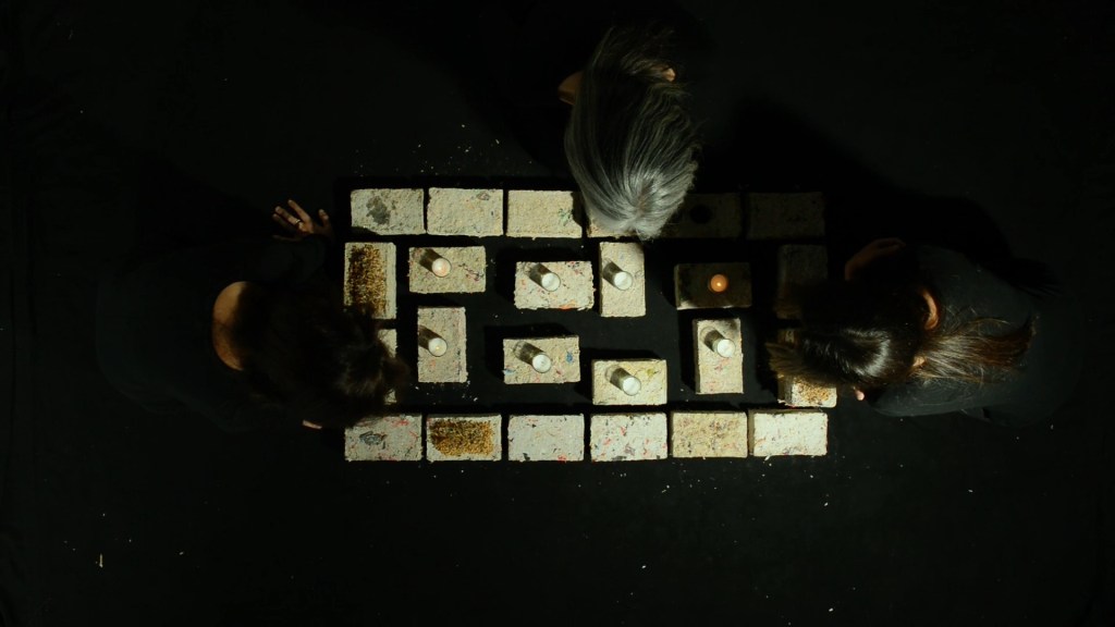 Still from Exile Echoes (2024) by Mazyar Mahan: women light candles around a brick memorial, seen from above