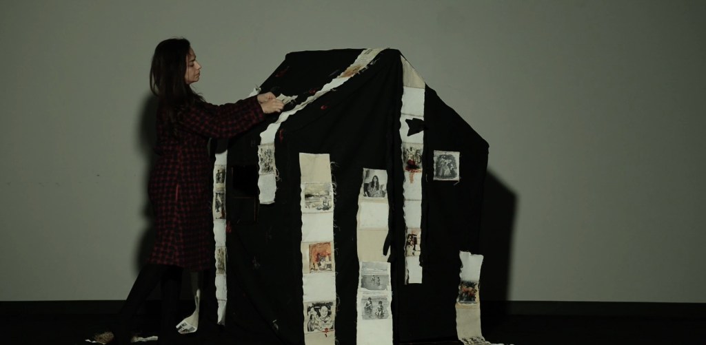 Still from Exile Echoes (2024) by Mazyar Mahan: a woman rests against the black fabric house stitched with photographs.