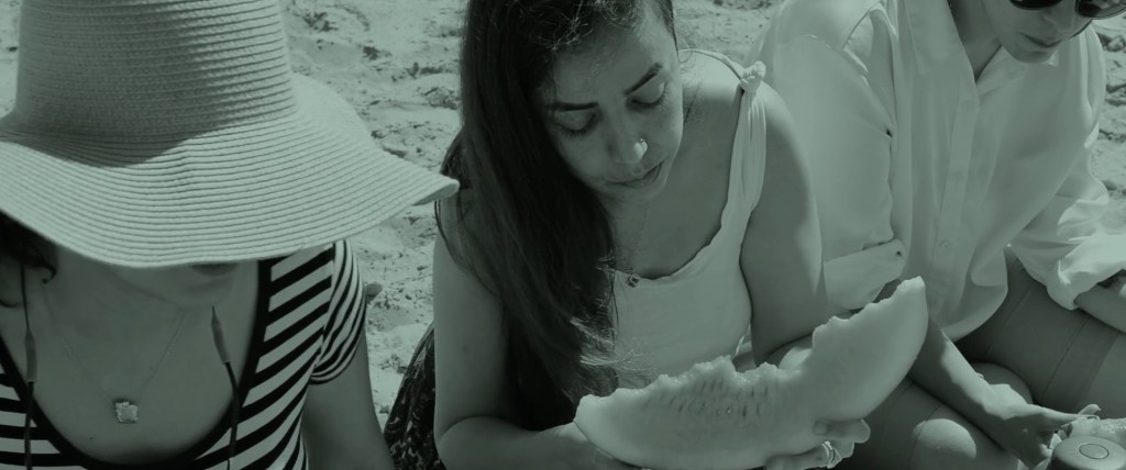 Still from Exile Echoes (2024) by Mazyar Mahan: three women sit together on the sand sharing watermelon, a memory from childhood.