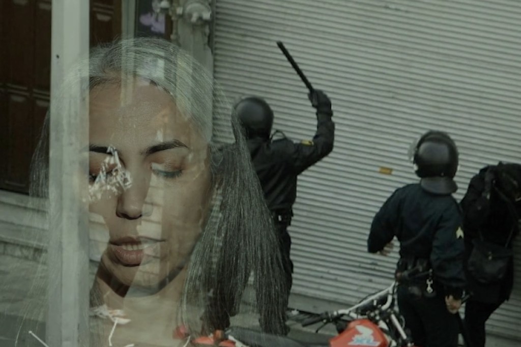 Still from Exile Echoes (2024) by Mazyar Mahan: archival footage of riot police raising a baton in a street in Iran, layered with a woman's face.